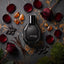 A faceted black perfume bottle labeled 'VIP BLEND OUD' sits on a dark stone surface surrounded by red rose petals, amber stones, dried pods, vanilla sticks, moss, and a red rose.